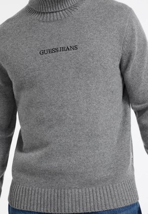 Gray knit sweater with a ribbed turtleneck and bottom hem. Features black embroidered "GUESS JEANS" logo on the chest. Smooth texture.