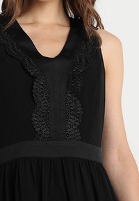 Černé šaty s výstřihem do V, vysoce detailní krajkovou výzdobou na přední straně a přiléhavým pasem s volnou sukní.