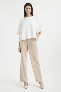 Maglione bianco oversize con maniche arrotolate, abbinato a pantaloni beige a gamba larga. Il modello indossa tacchi nudi e orecchini a cerchio dorati.