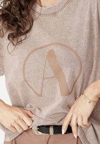 Brązowy, oversize T-shirt z wyblakłym logo "A" w jasnobrązowym kolorze. Miękki, teksturowany materiał. Czarny pasek z ozdobną srebrną klamrą. Prosty, casualowy design.