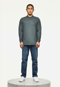 Mladý muž stojící na kulaté platformě, oblečený v šedém dlouhém polo tričku, modrých džínech a bílých teniskách, na jednobarevném bílém pozadí.