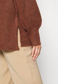 Sweter z dzianiny w kolorze rdzy o luźnym kroju, z bocznym rozcięciem i ściągaczami na rękawach. W połączeniu z jasnobeżowymi spodniami o szerokich nogawkach.