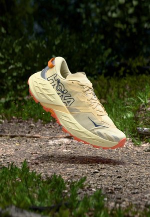 Beež ja oranž Hoka Speedgoat 7 maastikujooksu jalanõu õhus kivise pinnase kohal rohelise taustaga.