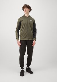 Maslinzasta zip-up jakna s crnim rukavima, teksturirana tkanina i logotip; uparen s crnim hlačama i crnim sportskim cipelama.