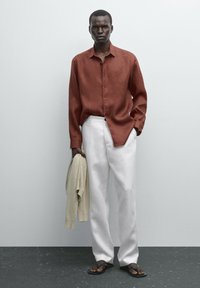 Red linen long-sleeve shirt with buttons, paired with white wide-leg trousers. The model holds a beige sweater and wears brown flip-flops.