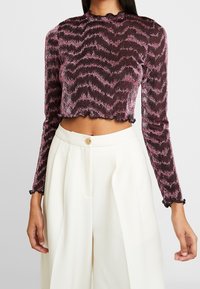 Långärmad, kort topp i mörkbrun med rosa zigzag-mönster. Volangkant och manschetter, med hög hals och strukturerad tyg.