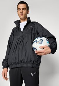 Vyras juodais sportiniais drabužiais laiko baltą ir mėlyną Nike futbolo kamuolį prieš vienodą foną.