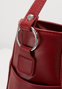 Sac à main en simili cuir rouge avec une attache ronde en métal, une texture lisse et une bandoulière sécurisée par un bouton-pression.