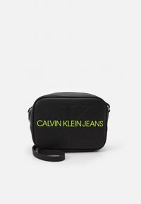 Czarna prostokątna torba wykonana z materiału syntetycznego, z wytłoczonym logo i jas Зеленым napisem "CALVIN KLEIN JEANS". W zestawie regulowany pasek.