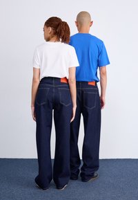 Unisex džinsai tamsiai mėlynos spalvos, su aukštu liemeniu, plačiomis kojomis ir oranžiniais logotipais ant nugaros. Derinkite su baltais ir mėlynais marškinėliais.
