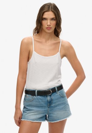 Top de algodón blanco sin mangas con tirantes finos, combinado con pantalones cortos de mezclilla azul claro con dobladillos deshilachados. Un cinturón de cuero negro acentúa el conjunto.