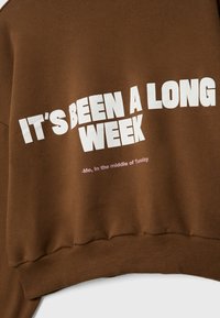 Sweat marron avec texte blanc indiquant "ÇA A ÉTÉ UNE LONGUE SEMAINE" et texte rose plus petit disant "-Moi, au milieu de mardi."