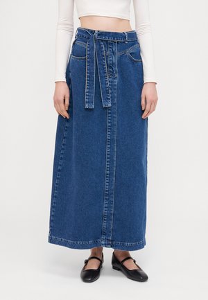 Femme portant une jupe en jean taille haute, longueur cheville, avec une ceinture, un crop top blanc à manches longues et des chaussures Mary Jane noires.