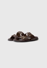 Καφέ δερμάτινα πέδιλα με δύο φαρδιές λωρίδες και χρυσούς μεταλλικούς αγκώνα, επίπεδη σόλα και λείο υλικό.