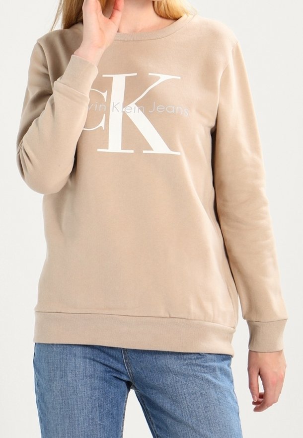 Felpa beige con scollo tondo, maniche lunghe e orlo a costine. Presenta un logo bianco prominente "CK" e la scritta "Calvin Klein Jeans."