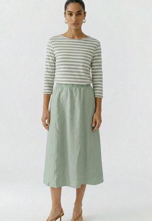 Femme portant une jupe midi vert clair et un haut à manches longues rayé gris et blanc, debout devant un fond blanc uni.