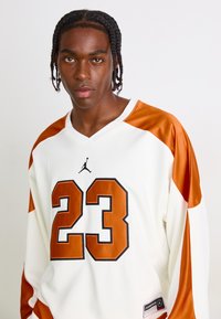 Young man with braided hair wearing white and orange basketball jersey numbered 23 with Jumpman logo on plain background.
