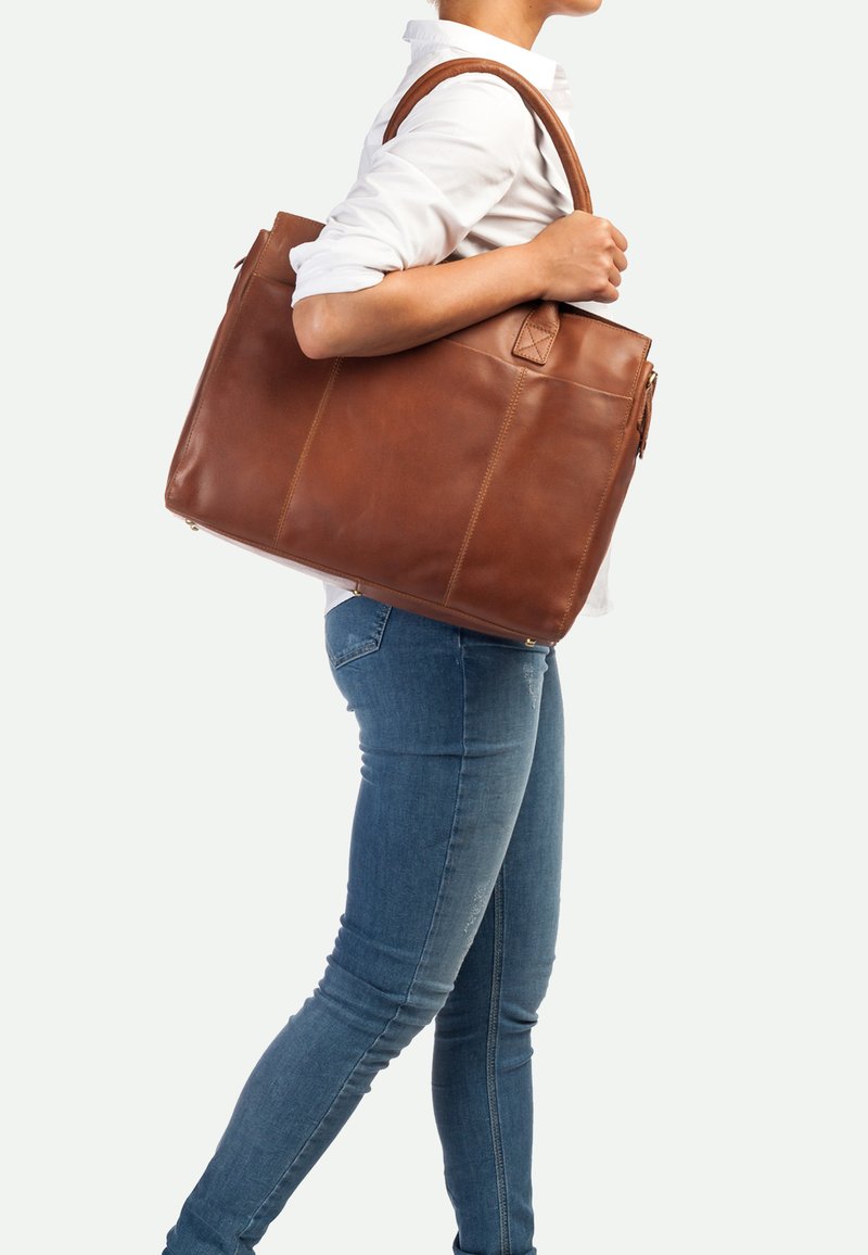 Braune Leder-Tote mit doppelten oberen Tragegriffen, glatter Textur, rechteckiger Form und sichtbaren Steppnähten. Über der Schulter getragen.