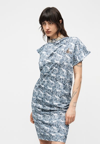 Γυναίκα με σκούρα μαλλιά που φοράει ένα μπλε φόρεμα με σχέδιο από αλογάκια-κουνιστά και μία πέρλα στο λαιμό, στέκεται μπροστά σε ένα απλό λευκό φόντο.