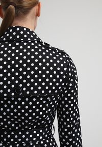 Černo-bílý trenčkot s puntíky, má límec, zapínání na knoflíky vpředu, pásek v pase a texturovanou tkaninu. Pohled ukazuje zadní design.