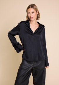 Svart satengbluse med V-hals, lange ermer og oversized mansjetter. Glatt tekstur, avslappet passform, kombinert med høyhåndte svarte bukser.