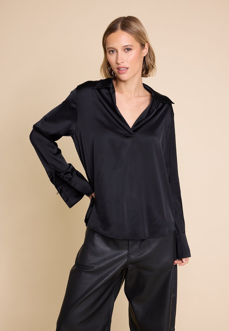 Svart satengbluse med V-hals, lange ermer og oversized mansjetter. Glatt tekstur, avslappet passform, kombinert med høyhåndte svarte bukser.