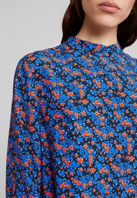 Chemise à motif floral en tissu bleu foncé, ornée de roses rouges et bleues. Col montant et manches longues, avec une texture lisse.