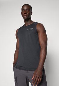 Black sleeveless athletic shirt made of textured fabric, featuring a logo on the upper left side, paired with gray pants.