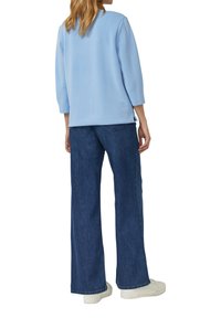 Jasnoniebieska bluza z krótkimi rękawami sięgającymi do połowy przedramienia, zestawiona z szerokimi ciemnymi dżinsami i białymi tenisówkami, widziana od tyłu.