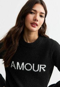 Zwarte trui van zacht materiaal met een ronde halslijn, met het woord "AMOUR" in witte vetgedrukte letters op de borst.