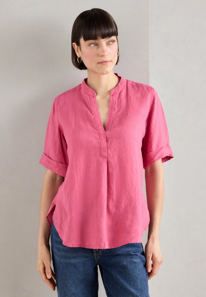 Blusa rosa in lino a maniche corte con scollo a V e risvolti, caratterizzata da spacchi laterali e una vestibilità comoda, indossata con jeans blu.