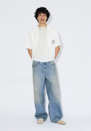 Camicia bianca a maniche corte sopra una t-shirt grigio chiaro, abbinata a jeans di denim blu slavato e larghi con lievi macchie e lavaggio chiaro.