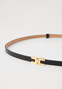 Černý kožený pásek s úzkým designem a zlatými doplňky. Disponuje charakteristickou zlatou sponou s geometrickým tvarem.
