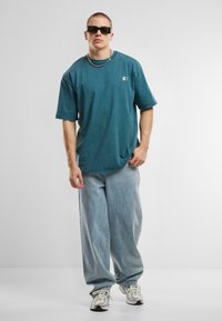 T-shirt en coton teal oversize à manches courtes, associée à un jean baggy en denim bleu clair. Les accessoires comprennent des lunettes de soleil noires et une chaîne en argent.