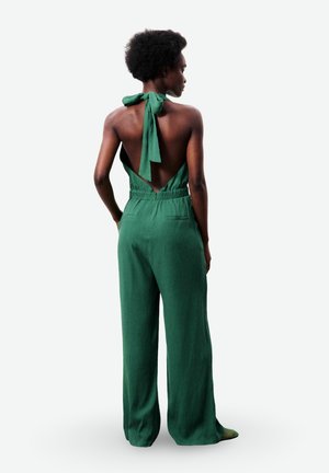 Femme aux cheveux courts et bouclés portant une combinaison verte sans manches avec un dos ouvert noué au niveau du cou, debout devant un fond blanc.