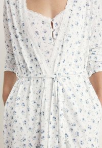 Robe florale en tissu blanc avec des motifs bleus, dotée de finitions en dentelle et d'une taille nouée. Le détail en boutons au niveau du col ajoute une touche délicate.