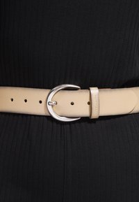 Cinturón de cuero beige con hebilla redonda plateada sujeto alrededor de la cintura sobre ropa de tela negra con relieve.