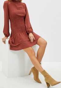 Rötlich-braues Kleid mit geraffter Taille, langen Puffärmel und geschichtetem Saum. Kombiniert mit beigefarbenen, schmal zulaufenden Stiefeletten mit blockabsatz.