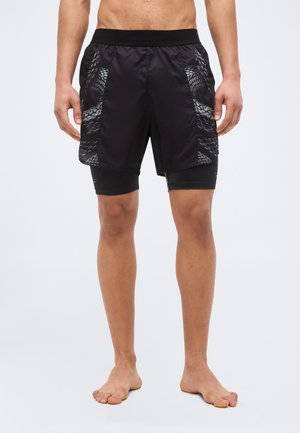 Άνδρας μοντέλο που φοράει μαύρα αθλητικά σορτς δύο σε ένα με πλαϊνές επιφάνειες σε σχήμα φιδιού, στέκεται ξυπόλητος σε απλό φόντο.