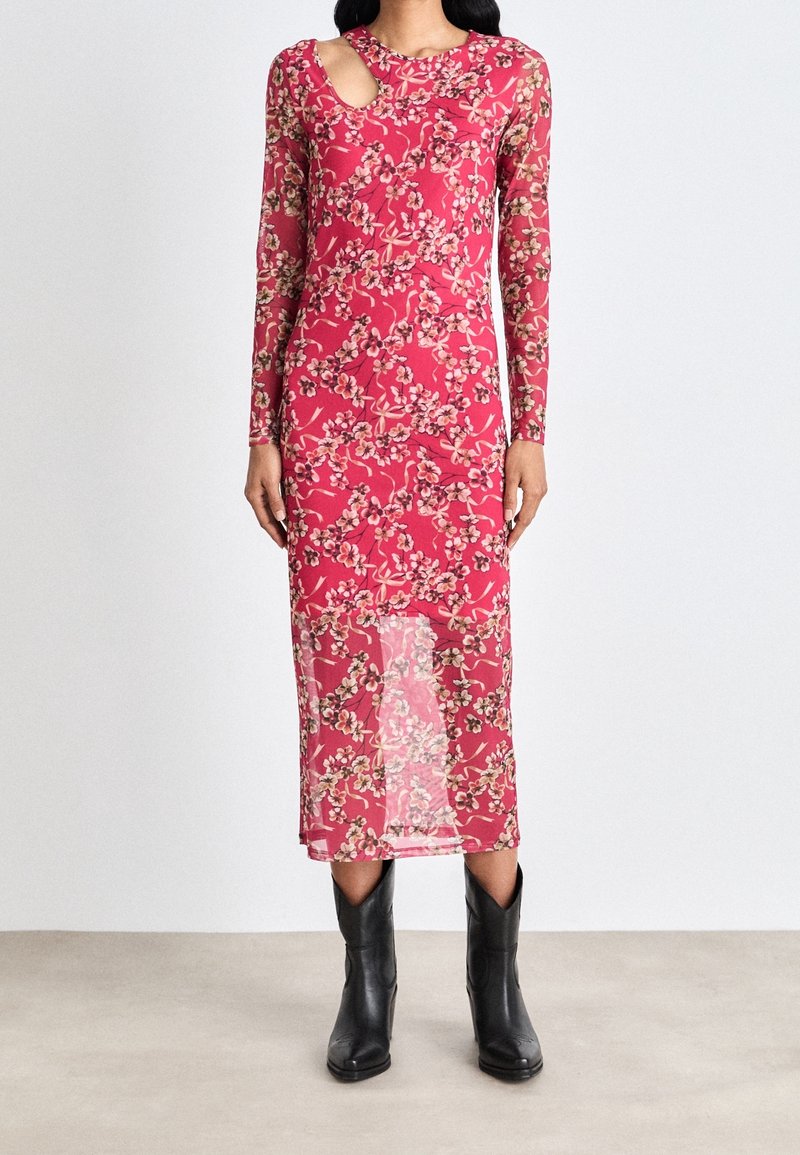 Femme portant une robe midi rose à fleurs à manches longues avec une découpe à l'épaule, associée à des bottes en cuir noires mi-mollet, sur un fond uni.