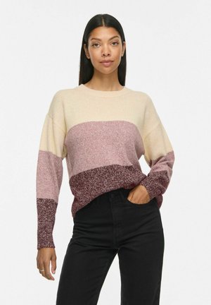 Femme aux longs cheveux foncés porte un pull color-block crème, rose et bordeaux, associé à un pantalon noir taille haute, debout avec une main dans la poche.