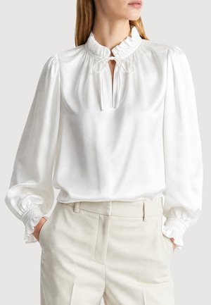 Femme portant un chemisier en satin blanc avec col à volants, encolure à nouer, manches longues bouffantes avec poignets froncés et pantalon taille haute beige clair.