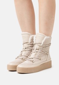 Beige vinterstøvler lavet af kunstig pels og læder, der har en tekstureret overflade, flad platformssål og rund tå med snøredetaljer.