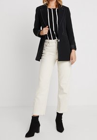 Kvinna i svart kavaj, svartvit randig topp, off-white avkortade jeans och svarta ankelstövlar, stående mot en enkel bakgrund.