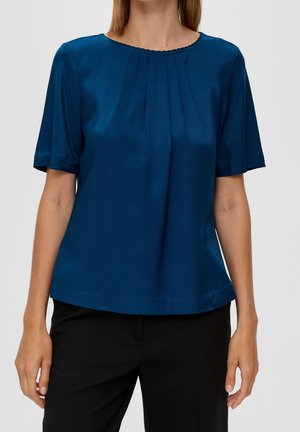 Nainen, jolla on lyhythihainen tummansininen pusero, jossa on laskoksia pääntiellä, ja mustat housut, seisoo yksivärisen taustan edessä.