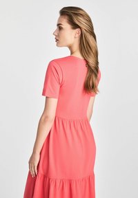 Korallowa sukienka z krótkimi rękawami, zebranym w pasie i zwiewnym dołem. Gładka tekstura z okrągłym dekoltem i detalem w postaci guzików z tyłu.