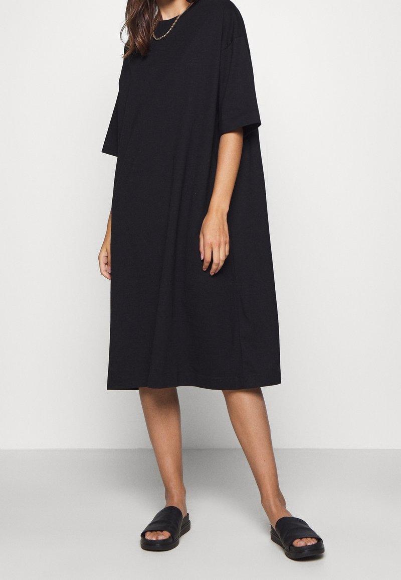 Robe t-shirt noire, surdimensionnée, à manches courtes, fabriquée en coton doux. Associée à des sandales noires à enfiler, avec une semelle plate.