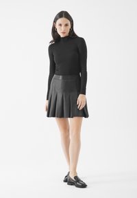 Donna con maglione a collo alto nero a maniche lunghe, gonna plissettata in pelle nera, mocassini neri, in piedi contro uno sfondo bianco.