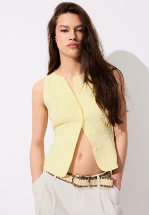 Jeune femme aux longs cheveux bruns porte un gilet jaune sans manches boutonné et un pantalon blanc taille haute avec une ceinture beige, debout contre un mur blanc.