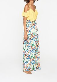 Top giallo scoperto sulle spalle con volant, abbinato a una gonna maxi con stampe geometriche multicolori. Sandali leggeri.